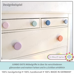 Op de afbeelding: Een witte commode met vier lades en kleurrijke ronde knoppen. De knoppen zijn in tinten roze, blauw, lichtblauw en perzik. De commode is gelabeld "JUMBO DOTS Möbelgriffe in über 80 verschiedenen glänzenden und matten Farben und in 4 Größen erhältlich 100% handgefertigt 100% handbemalt 100% MADE IN GERMANY".