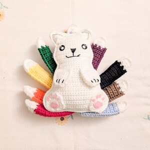 May include: A white crocheted fox plush toy with nine colorful tails in green, purple, black, brown, light blue, red, orange, and yellow. The fox features embroidered black eyes, a nose, a mouth, and light pink felt paw pads on its feet.
