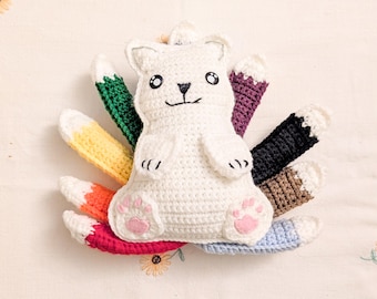 May include: A white crocheted fox plush toy with nine colourful tails in green, purple, black, brown, light blue, red, orange, and yellow. The fox features embroidered black eyes, a nose, a mouth, and light pink felt paw pads on its feet.