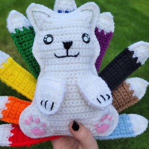 May include: A white crocheted cat with rainbow colored tails. The cat has black eyes and a black nose. The tails are crocheted in different colors of the rainbow.