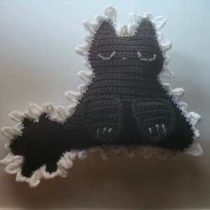 Puede incluir: Un peluche de gato hecho a mano en crochet negro con detalles blancos. El gato tiene los ojos cerrados y detalles bordados en blanco. Los bordes están adornados con un borde blanco con volantes. El gato está sentado con la cola extendida.