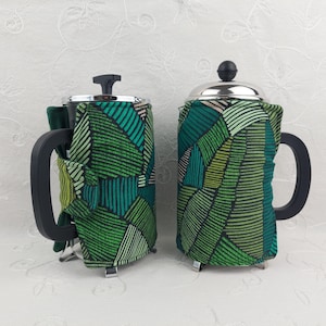 May include: Two French press coffee makers with black handles and stainless steel lids. Each is wrapped in a green fabric with a geometric pattern of lines and shapes in shades of green and beige. The coffee makers sit on a white surface.