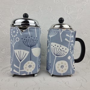 May include: Two French press coffee makers with stainless steel tops and black handles. Each is wrapped in a light blue fabric with a white and navy floral pattern. The coffee makers are on a white surface.