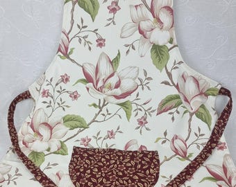 Könnte beinhalten: Eine Schürze mit cremefarbenem Hintergrund und einem Blumenmuster aus großen rosa-weißen Magnolienblüten und grünen Blättern. Die Schürze verfügt über eine dunkel kastanienbraune Tasche mit kleinem Blumenmuster und passende Riemen.