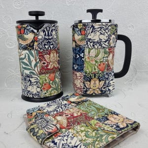 May include: A French press, a coffee mug, and a small case are displayed. Each item is covered in a patchwork fabric with a floral and bird pattern in shades of blue, red, green, and cream. The French press and mug have black handles and lids.