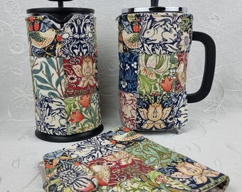 Könnte beinhalten: Zwei French-Press-Kaffeebereiter und eine passende rechteckige Stoffhülle, alle mit einem Patchwork-Design aus Blumen- und Hasenmotiven in Marineblau, Grün, Rot und Creme. Jeder Kaffeebereiter hat einen schwarzen Deckel, Griff und Boden.