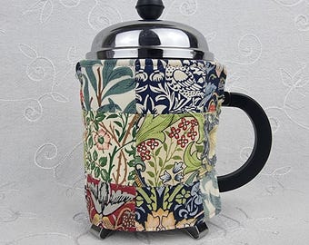 Könnte beinhalten: Eine French-Press-Kaffeebereiter mit einem Patchwork-Stoffüberzug, der verschiedene Blumen- und Vogelmuster in Marineblau, Creme, Grün und Rot aufweist. Der Kaffeebereiter hat einen Deckel aus poliertem Chrom mit schwarzem Knopf und Griff.
