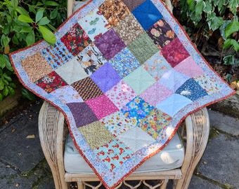 Könnte beinhalten: Eine quadratische Patchwork-Decke aus verschiedenen Stoffquadraten in unterschiedlichen Farben und Mustern, darunter Blumen-, geometrische und Tiermotive. Die Decke ist mit einem schmalen, orange-roten Rand versehen und weist sichtbare Steppnähte auf.