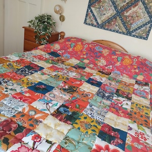 May include: A colorful patchwork quilt with a floral pattern, featuring squares of various colors including red, orange, blue, and yellow. The quilt is on a bed with a red floral patterned headboard. A small wall hanging with a floral design is above the bed.