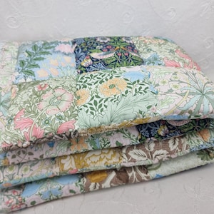 May include: A stack of quilted fabric squares in various floral patterns. The squares feature a mix of colors, including pink, blue, green, and brown, with detailed botanical designs. The fabric appears soft and padded, suggesting a handmade item.
