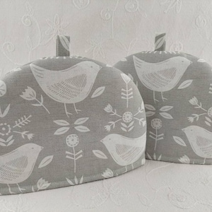 Peut inclure: Deux couvre-théières gris avec un motif d'oiseaux et de fleurs blancs. Chaque couvre-théière a une petite boucle en haut. Le motif d'oiseau est répété sur le tissu, créant un aspect charmant. Les couvre-théières sont conçus pour garder le thé au chaud.