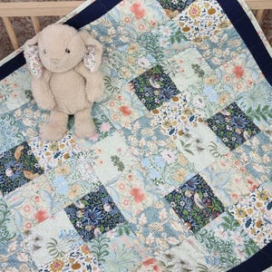 May include: A beige plush rabbit toy with floral-lined ears sits on a patchwork quilt. The quilt features a repeating pattern of botanical illustrations in shades of blue, green, peach, and yellow, finished with a solid dark navy blue border.