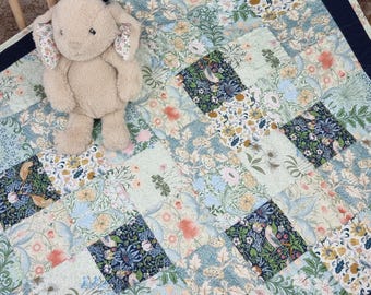 Könnte beinhalten: Ein beiges Plüschhasenspielzeug mit blumig gemusterten Ohren liegt auf einer Patchwork-Decke. Die Decke zeigt ein wiederkehrendes Muster aus botanischen Illustrationen in Blau-, Grün-, Pfirsich- und Gelbtönen mit einem dunkelblauen Rand.