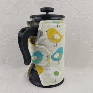 May include: A French press coffee maker with a decorative cozy. The cozy is cream-colored and features a repeating pattern of colorful birds in yellow, blue, gray, and white. The press has a black handle and lid.