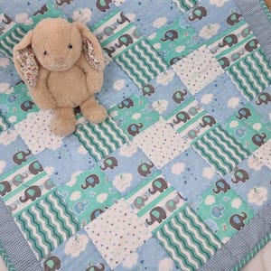 May include: A beige plush rabbit with floral-patterned inner ears resting on a patchwork quilt. The quilt features alternating squares of light blue and teal fabric decorated with grey and blue elephant illustrations, white clouds, and wavy lines.