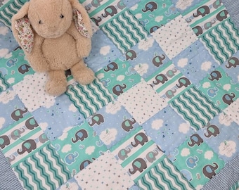 Könnte beinhalten: Ein beiger Plüschhase mit floral gemusterten Innenohren auf einer Patchwork-Decke. Die Decke besteht aus abwechselnden hellblauen und türkisfarbenen Stoffquadraten, die mit grauen und blauen Elefanten, weißen Wolken und Wellenlinien verziert sind.