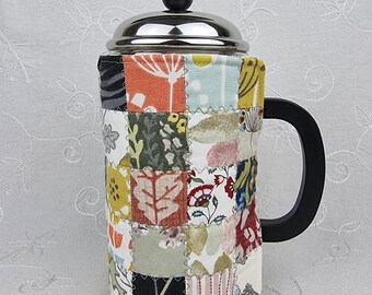 Könnte beinhalten: Eine French-Press-Kaffeebereiter mit einem Edelstahldeckel, einem schwarzen Knopf und einem schwarzen Griff. Die Glaskaraffe ist von einer gesteppten Stoffhülle umgeben, die aus Patchwork-Quadraten in verschiedenen Farben wie Orange, Gelb, Grün und Weiß besteht.