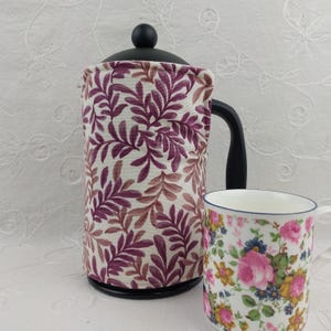 May include: A black French press with a purple and brown leaf-patterned cozy and a floral-decorated mug. The cozy covers the glass carafe, and the mug features pink roses and blue accents. The set is on a white surface.