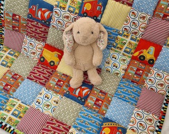 Könnte beinhalten: Eine Patchwork-Decke mit Quadraten mit Lastwagen, Feuerwehrautos und anderen Fahrzeugen in Rot, Blau, Gelb und Grün. Ein Plüsch-Hasenspielzeug sitzt auf der Decke. Der Rand der Decke hat ein schwarz-weißes Streifenmuster.