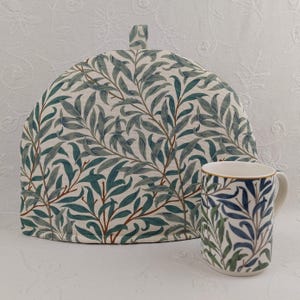 May include: A white tea cosy with a green and brown willow pattern design. A matching mug with a blue and green willow pattern design is in the foreground.