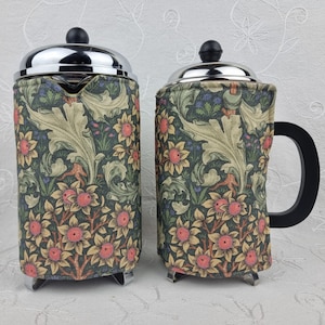 May include: Two stainless steel French press coffee makers with black handles and lids. The coffee makers are covered in a floral fabric with a pattern of pink flowers, green leaves, and a dark blue background. The coffee makers sit on silver metal feet.