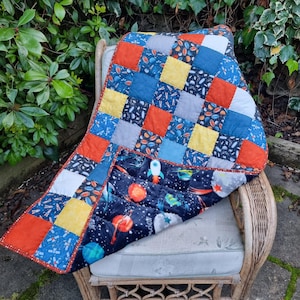 May include: A patchwork quilt featuring a grid of solid orange, yellow, blue, and grey squares alongside squares with a space-themed print. The bottom section displays a larger fabric panel with a dark blue background, colourful planets, and rockets.