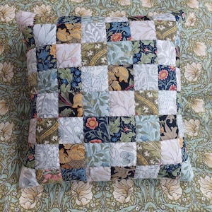 May include: A square patchwork cushion with a checkered pattern. The cushion is made of different fabrics with floral and geometric designs in shades of blue, green, yellow, and white.