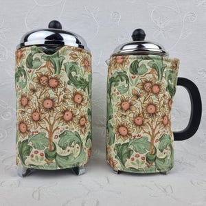 May include: Two French presses with floral patterned sleeves. The sleeves feature a cream background with a repeating pattern of sunflowers, green leaves, and brown stems. The presses have chrome lids and bases, and black handles.