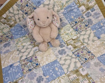 Könnte beinhalten: Ein beigefarbener Plüschhase mit langen Ohren und floralem Innenfutter sitzt auf einer Patchwork-Decke. Die Decke besteht aus quadratischen Stoffbahnen in Salbeigrün, Hellblau und Weiß, die verschiedene botanische und florale Muster aufweisen.