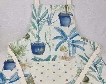 Könnte beinhalten: Eine Schürze mit einem botanischen Druck aus blauen Topfpflanzen, grünem Blattwerk und weißen Blüten auf cremefarbenem Grund. Die Schürze verfügt über eine halbkreisförmige Vordertasche mit einem Muster aus kleinen blauen und braunen Punkten.
