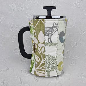 May include: A French press coffee maker featuring a fabric cosy with a patchwork design. The fabric displays illustrations of a fox, birds, and trees in shades of green, grey, and yellow. It has a black plastic handle and a chrome lid with a black knob.