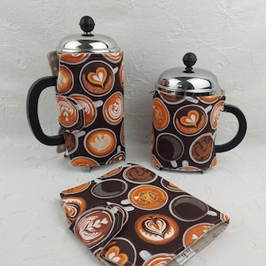 Peut inclure: Trois housses pour cafetières à piston et un dessous de verre assorti. Les housses et le dessous de verre présentent un fond marron avec des motifs de latte art orange, blanc et marron. Les cafetières ont des poignées noires et des couvercles argentés.