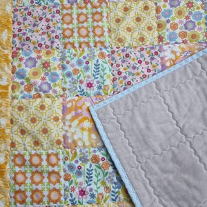 May include: A patchwork quilt featuring a variety of floral and geometric patterns in shades of yellow, pink, orange, blue, and green. One side displays a grid of colorful fabric squares, while the other side is a solid, quilted light gray fabric.