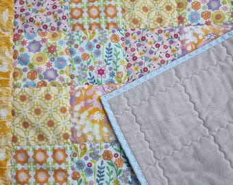 Könnte beinhalten: Eine Patchwork-Decke mit verschiedenen Blumen- und geometrischen Mustern in Gelb-, Rosa-, Orange-, Blau- und Grüntönen. Eine Seite zeigt ein Gitter aus bunten Stoffquadraten, während die andere Seite aus einem einfarbigen, hellgrauen Steppstoff besteht.
