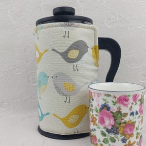 May include: A French press coffee maker with a bird-patterned cozy and a floral-patterned mug. The cozy features gray, yellow, and blue birds on a light gray background. The mug has pink roses and blue flowers.