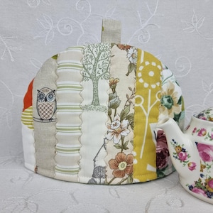 May include: A quilted fabric tea cozy featuring vertical panels with diverse patterns including a brown owl, green stripes, a tree silhouette, floral motifs in orange and yellow, and a white teapot with pink and green floral patterns beside it.