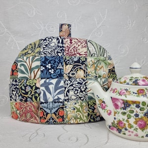 May include: A patchwork tea cosy featuring a variety of floral and bird patterns in blue, green, red, and cream. A white teapot with a floral design is next to the cosy.