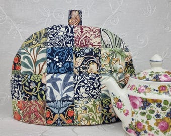 Könnte beinhalten: Ein Patchwork-Teewärmer mit verschiedenen Blumen- und Vogelmustern in Blau-, Grün-, Rot- und Cremetönen. Eine weiße Teekanne mit floralem Design steht daneben.