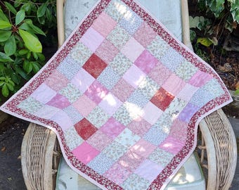 Könnte beinhalten: Ein quadratischer Patchwork-Quilt mit einem Gitter aus abwechselnden Stoffquadraten in verschiedenen Rosa-, Hellrot- und Weißtönen, wobei einige Quadrate florale Muster aufweisen. Der Quilt ist mit einer dunkelroten Bordüre mit weißem Blumenmuster eingefasst.