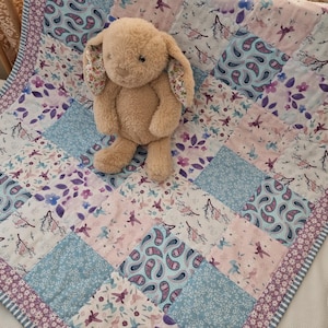 May include: A tan plush bunny sits on a patchwork quilt. The quilt features a mix of floral, paisley, and bird patterns in shades of blue, pink, and purple. The quilt's border has a floral design.