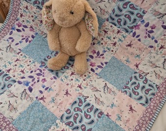 Könnte beinhalten: Ein beiges Plüschkaninchen liegt auf einer Patchwork-Decke. Die Decke zeigt Blumen-, Paisley- und Vogelmuster in Blau-, Rosa- und Lilatönen. Der Rand der Decke hat ein Blumenmuster.