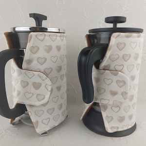 May include: Two French press coffee makers with heart-patterned cozies. The cozies are white with a repeating pattern of light brown hearts. The coffee makers have black handles and lids. The glass carafes are visible through the cozy openings.