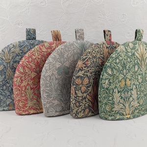 May include: Five tea cosies in a row, each with a different floral pattern. The cosies are in shades of blue, red, grey, dark green, and light green. Each cosy has a small loop at the top.