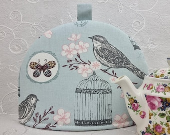Könnte beinhalten: Hellblaue Teewärmer mit Vogel- und Blumenmuster. Das Design zeigt einen Vogel auf einem Ast mit rosa Blüten, einen Schmetterling und einen Vogelkäfig. Eine weiße Teekanne mit floralem Muster ist teilweise sichtbar.