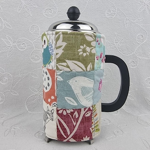 May include: A French press coffee maker with a patchwork fabric cosy. The cosy features various fabric squares with floral, bird, and owl patterns in shades of green, blue, orange, and red. The handle is black, and the lid is silver.
