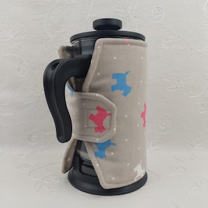 May include: A French press coffee maker with a beige insulated cozy. The cozy features a pattern of pink and blue dog silhouettes and white polka dots. The handle and lid are black.