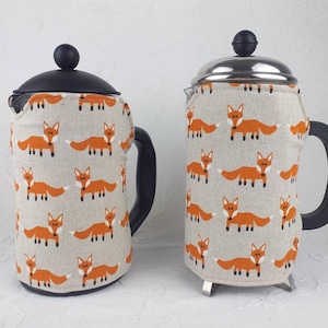 May include: Two coffee carafes with fox print fabric covers. The carafes are made of glass and have a stainless steel lid. The fabric covers are beige with orange foxes printed on them.