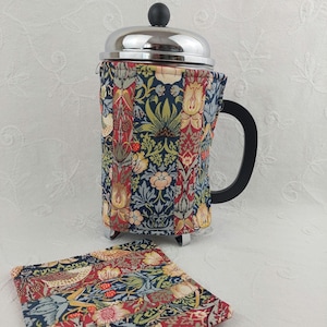 May include: A stainless steel French press coffee maker with a black handle and a decorative fabric cozy. The cozy features a floral pattern in shades of blue, red, and yellow. A matching coaster is also present.