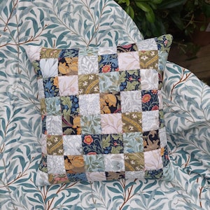 May include: A square patchwork cushion featuring a range of floral designs in shades of blue, green, yellow, pink, and white. The cushion is crafted from fabric squares stitched together to form a chequered pattern.