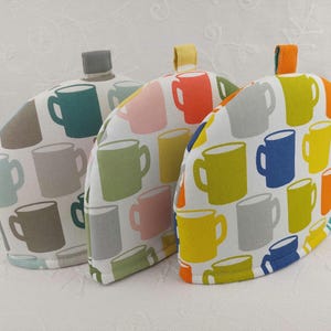 May include: Three tea cozy covers with a white background and colorful mugs printed on them. The covers are made of fabric and have a rounded shape. The mugs are in various colors, including blue, green, yellow, orange, and pink.